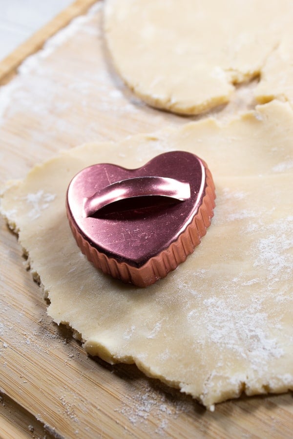 rolled out cream wafer dough on a bamboo cutting board with a copper heart cookie cutout on it 