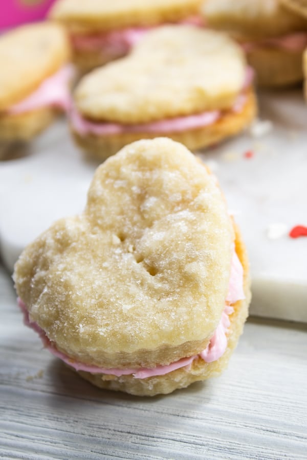 a white board with a heart shaped cream wafer cookie on it with red sprinkles and more heart shaped cookies in the background