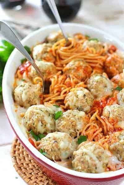 Meatballs and Spaghetti baked in a casserole dish, garnished with parsley and parmesan with forks spinning the pasta. 