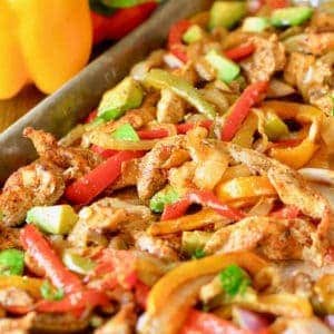 Sheet Pan Fajitas fresh out of the oven- sliced chicken, tried colored bell peppers and sliced onions on a baking sheet topped with avocado, red onion, and cilantro.