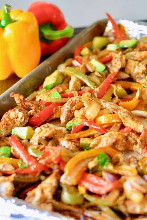 Sheet Pan Fajitas fresh out of the oven- sliced chicken, tried colored bell peppers and sliced onions on a baking sheet topped with avocado, red onion, and cilantro.