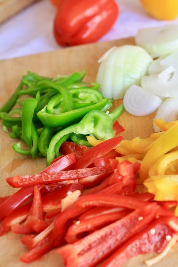 Sliced red, green and yellow bell peppers with sliced yellow onion on a cutting board.
