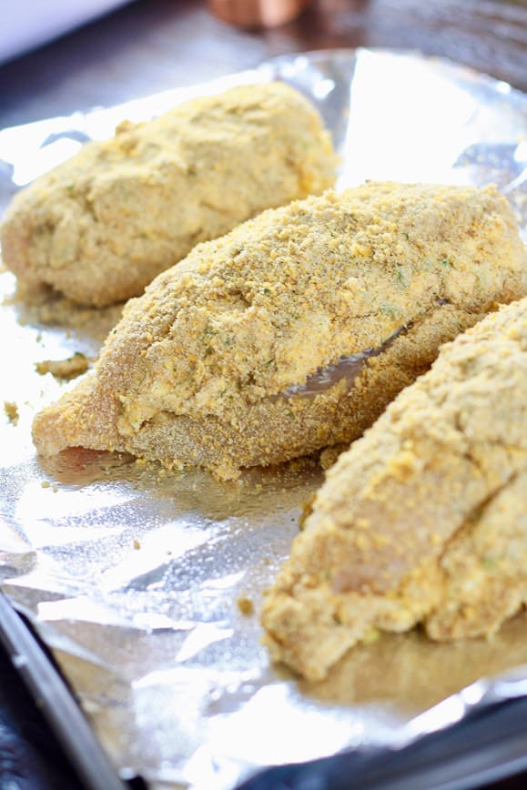 Chicken stuffed with jalapeño cream cheese & cheddar cheese mixture coated in egg mixture and bread crumbs on a baking sheet ready to go in the oven.