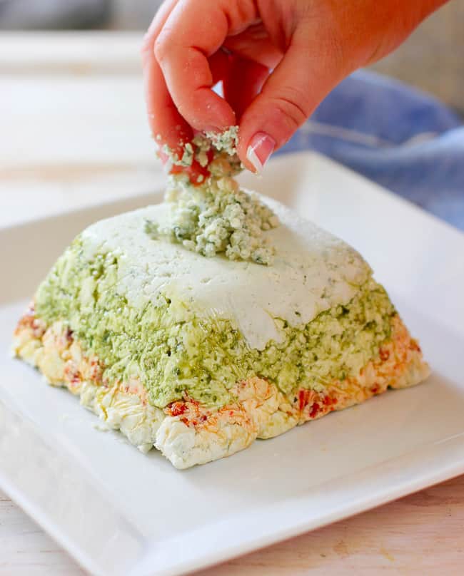 3 layer dip with pesto, sun-dried tomatoes, and blue cheese being toped with crumbled blue cheese