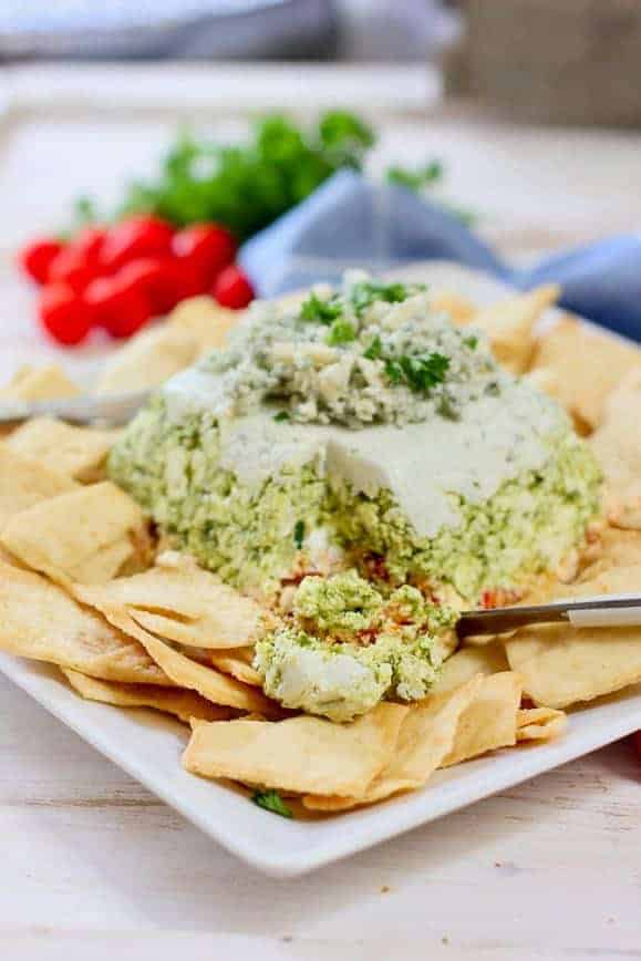 A 3 layer dip on a white platter surrounded by pita chips with a section cut out with a serving knife. 