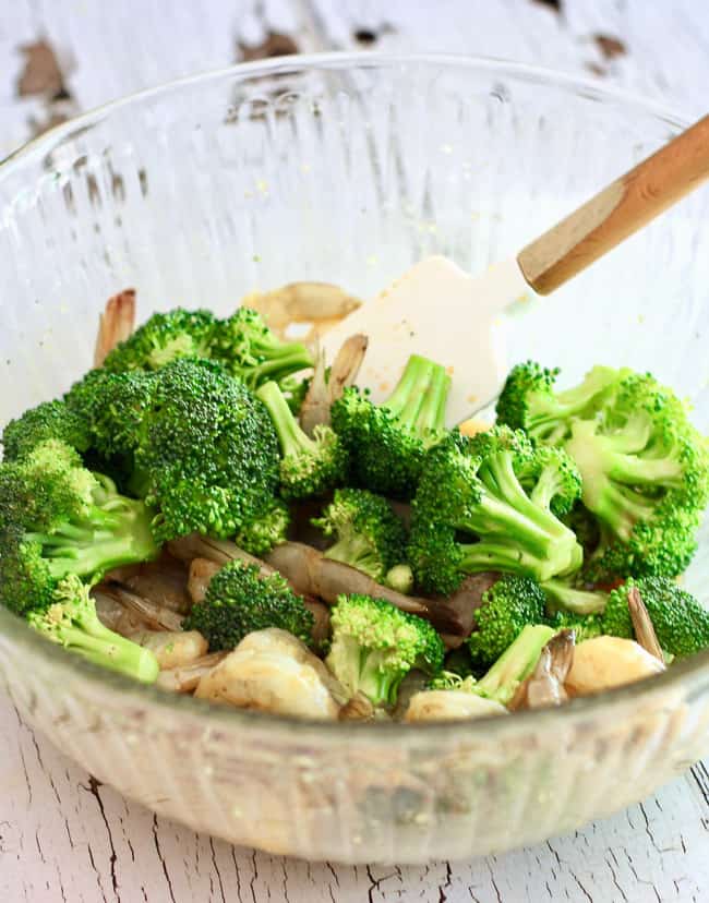 A clear bowl with a silicone spatula filled with broccoli, raw shrimp and stir fry marinade