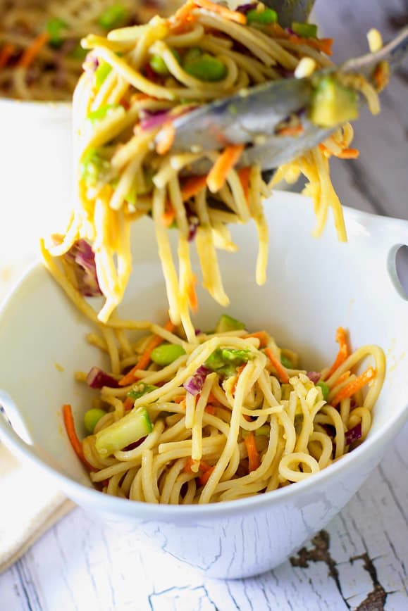 A white bowl of soba noodle salad being spooned out with silver tongs