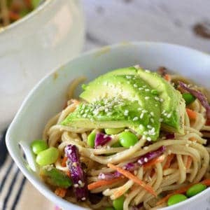 A white bowl filled with soba noodle salad and topped with avocado and sesame seeds