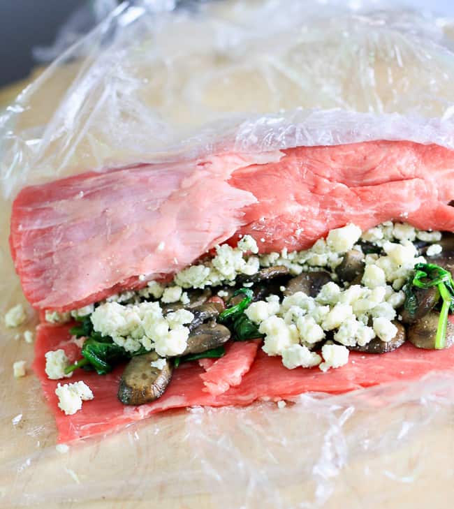 A flank steak roll stuffed with blue cheese, spinach and mushrooms being rolled on a cutting board with Saran Wrap