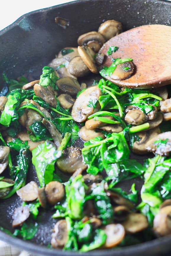 A cast iron pan filled with spinach and mushrooms being cooked in butter and garlic with a wooden spoon