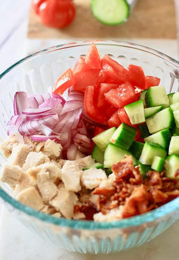 A glass bowl filled with chopped veggies, chicken, cucumbers, tomatoes, red onion and bacon