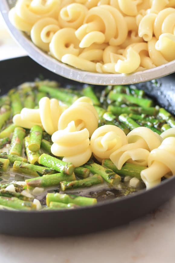 a skillet of asparagus and butter mixture with pasta being poured into it with a silver strainer