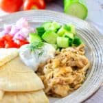 slow cooker greek chicken on a plate with cucumbers, red onion, tomato, pita and Tzatiki