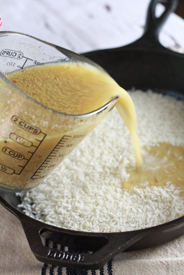 A black cast iron pan with uncooked white rice with a broth honey mustard mixture being poured over it