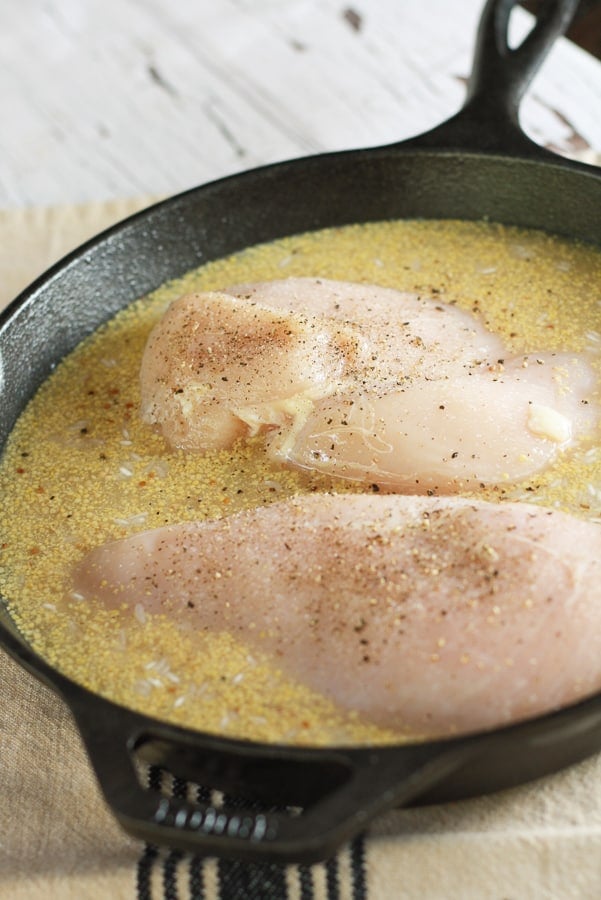 A black cast iron pan with a mixture of raw chicken, rice, and broth mixture sitting on a white board.