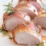 sliced proscuitto wrapped pork tenderloin on parchment paper with rosemary garnish