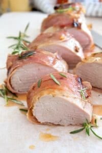 sliced proscuitto wrapped pork tenderloin on parchment paper with rosemary garnish