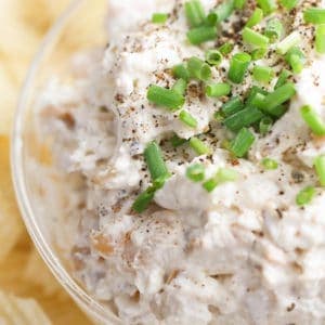 A clear bowl surrounded by chips, the clear bowl is filled with a creamy French onion dip mixture and garnished with fresh ground black pepper and chopped chives