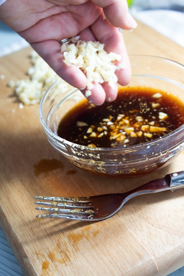 A clear bowl on a bamboo cutting board filled with soy marinade with chopped garlic being added in