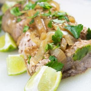 A pork loin on a white platter with slices of braised onion, fresh lime wedges and cilantro