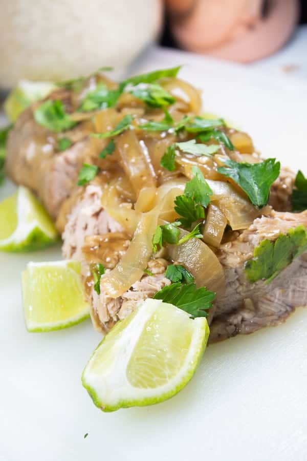 A pork loin on a white platter with slices of braised onion, fresh lime wedges and cilantro