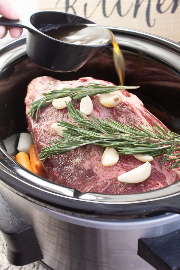 a raw pot roast in a slow cooker garnish with fresh rosemary sprigs and garlic