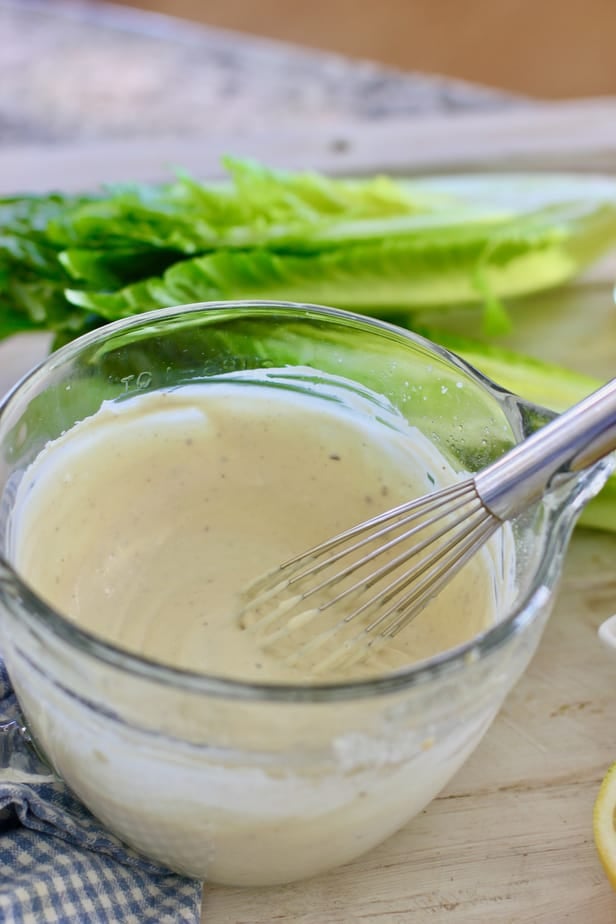 whisking a clear bowl of caesar dressing with romaine lettuce in the background