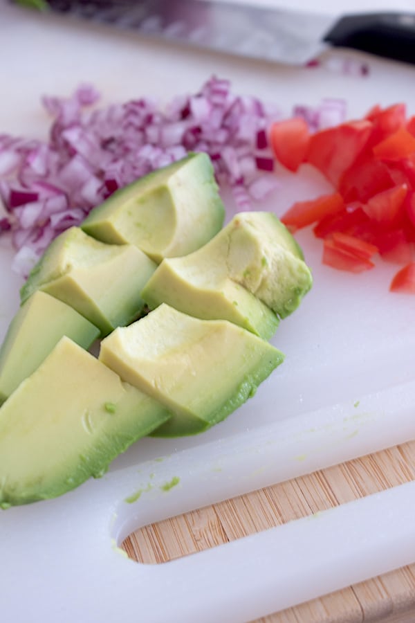 chunks of avocado and diced tomatoes and purple onion on a white cutting board 