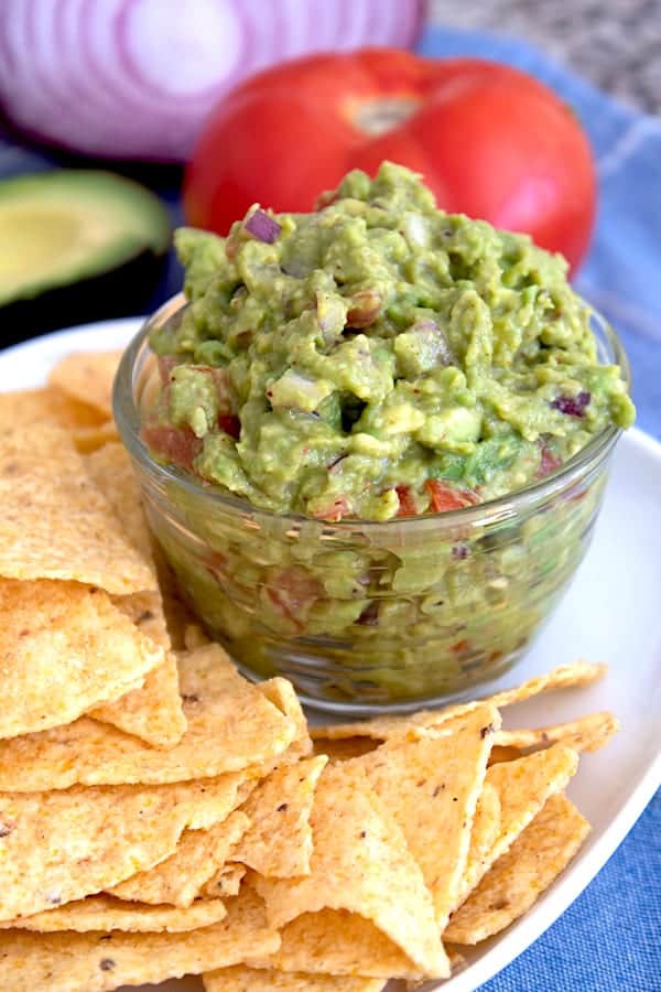 a clear bowl of guacamole on a white plate with chips and tomato and red onion in the background