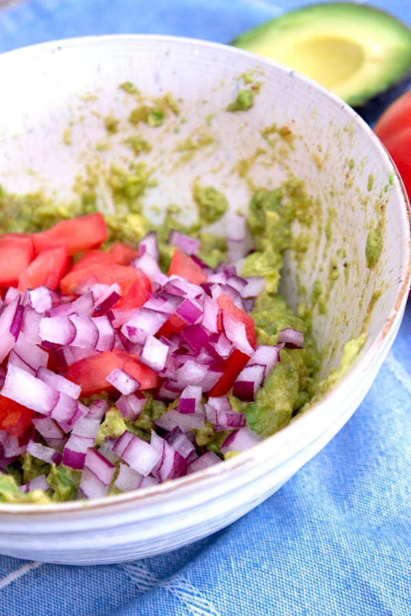 adding purple onion tomato to guacamole in a ceramic bowl on a blue linen towel