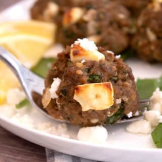 greek meatball with browned feta on a white plate with lemons and spinach in background