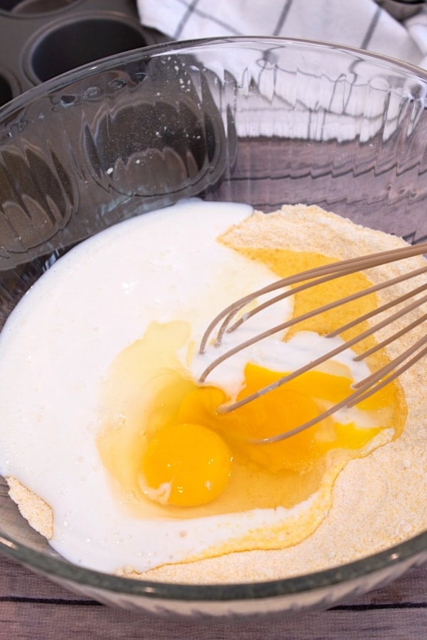 mixing ingredients for cornbread muffins in a clear bowl with a whisk
