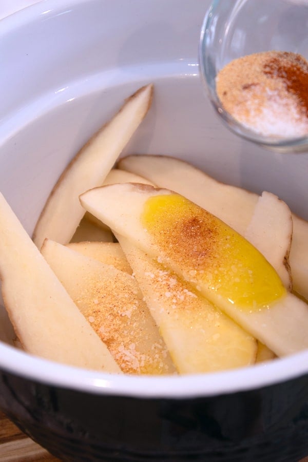 adding olive oil and spices to raw potato wedges in bowl