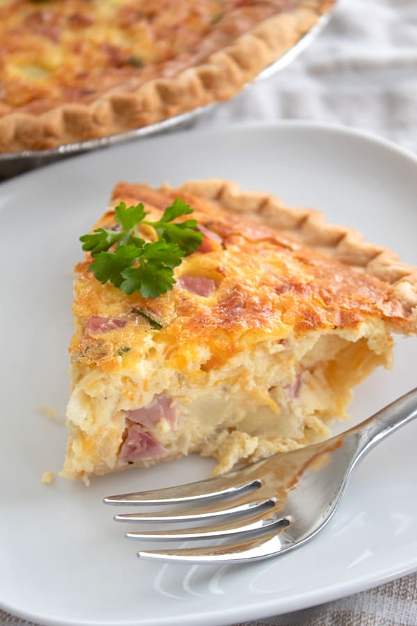 a slice of quiche on a white plate with a fork and parsley garnish