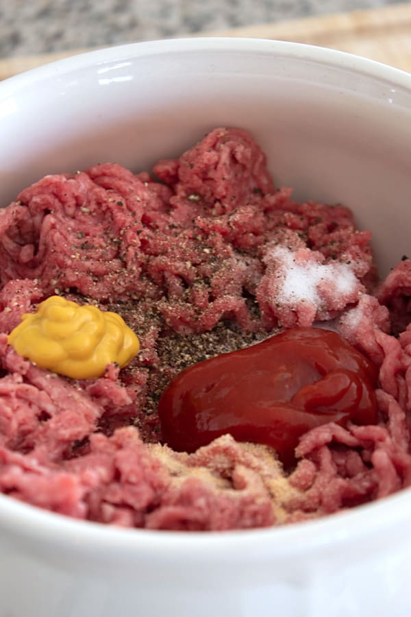 mixing ground beef in a white bowl with ketchup, mustard salt, onion powder and pepper