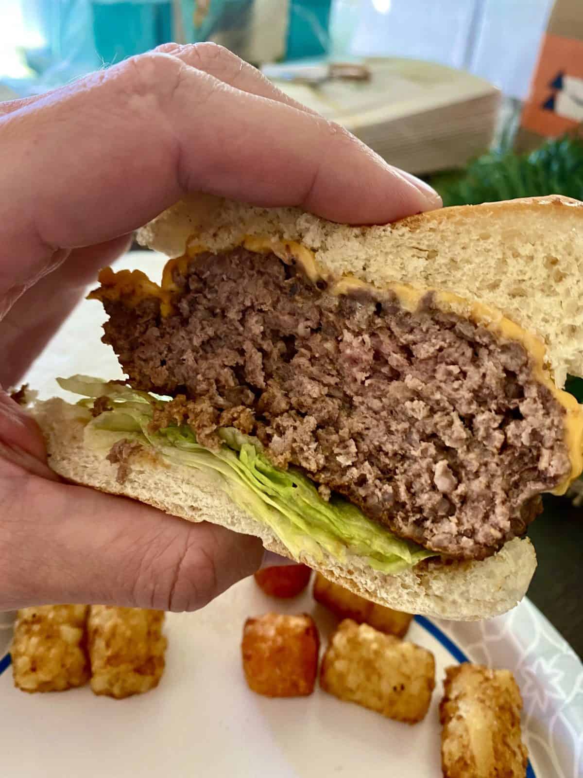 https://laughingspatula.com/wp-content/uploads/2021/12/holding-medium-well-burger-with-tater-tots-in-background-scaled.jpeg