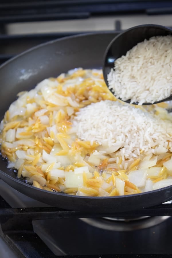 toasted orzo and onion in a pan with a scoop of rice going in