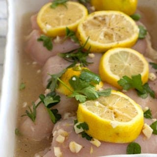 white casserole dish full of chicken topped with lemon slices and fresh herbs in marinade
