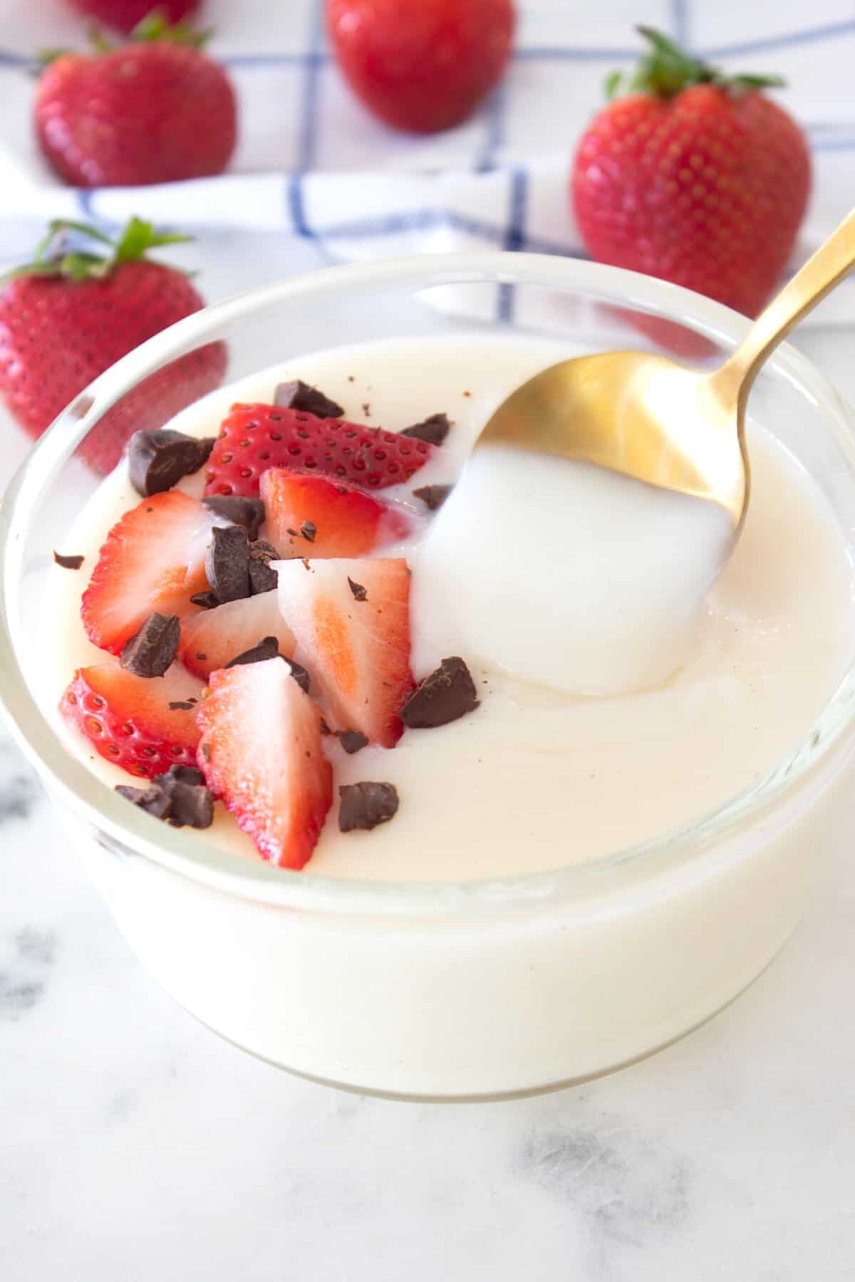 vanilla pudding in a clear dish with strawberries and chocolate garnish and a gold spoon spooning some
