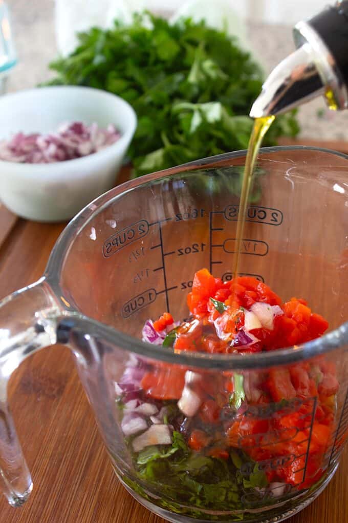 adding olive oil to dish full of herbs and roasted red peppers